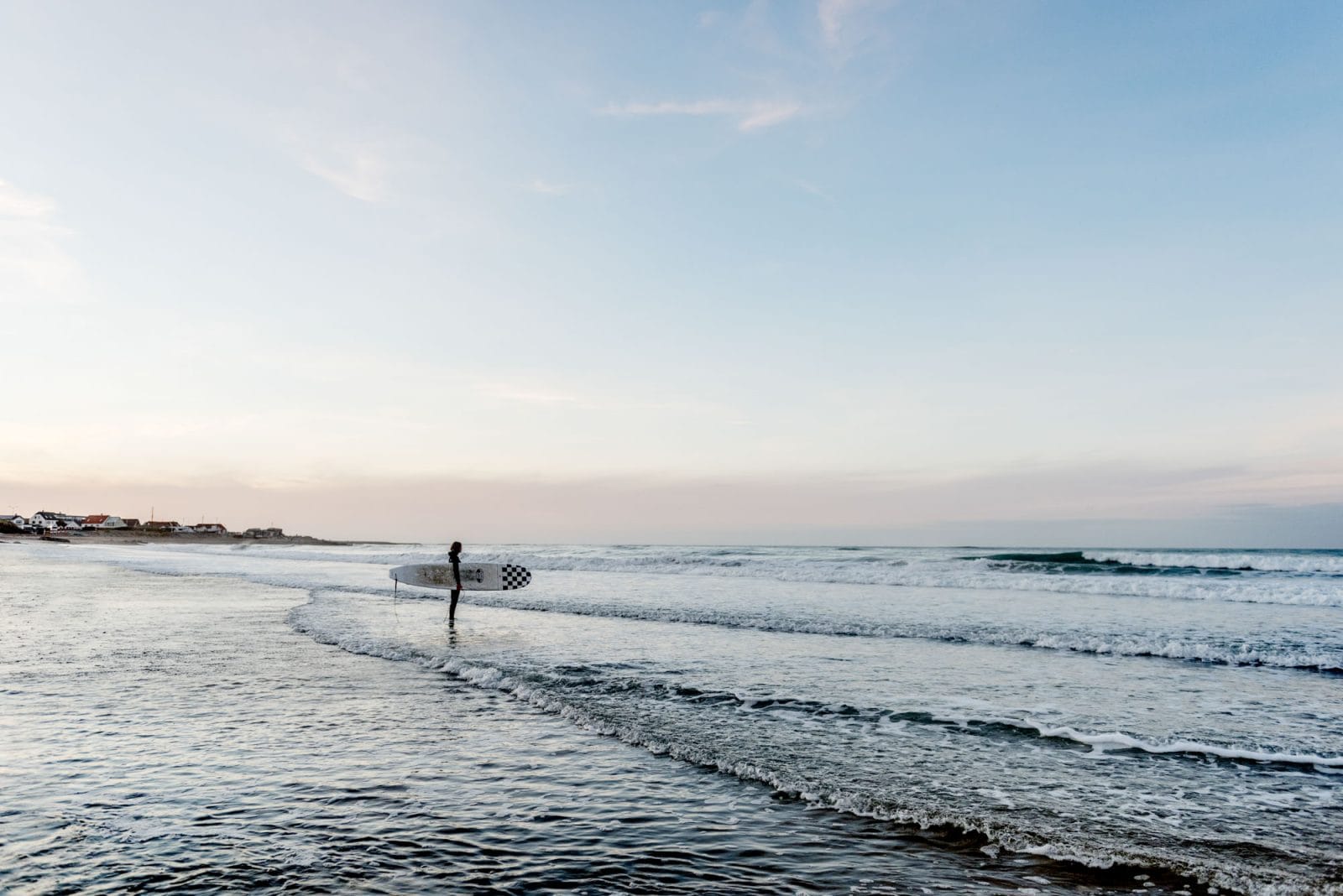 Surfer med surfboard står i vand ser mod bølgerne