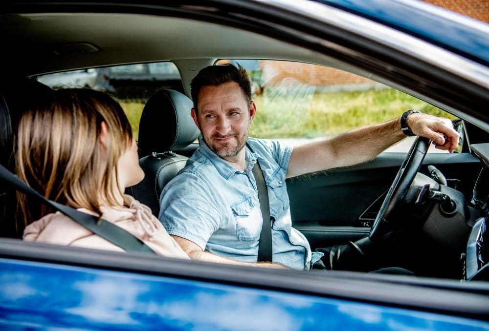 en mand og en pige sidder i en blå bil Manden er forsikret med en førerpladsdækning, så han også er dækket i tilfælde af uheld.